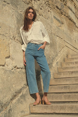 A standing woman wearing light blue denim jeans and white long sleeve blouse